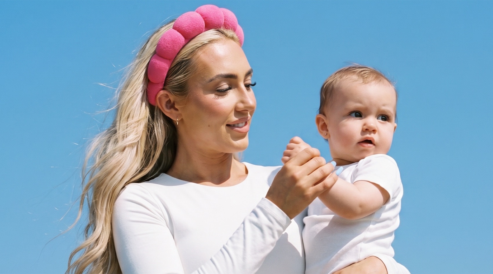 Polly lifestyle — a mother in a pink headband holding her baby against a blue sky